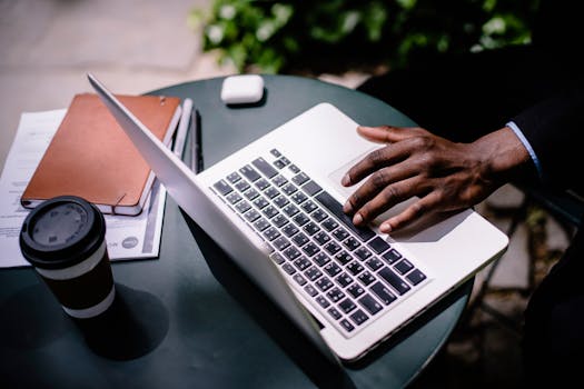 A businessman uses a laptop at an outdoor cafe table with coffee and documents nearby.