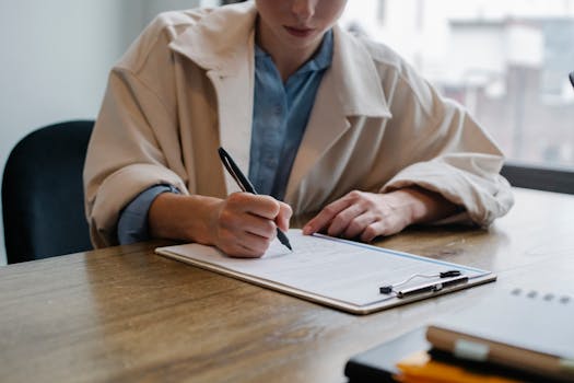 A woman in a formal setting fills out paperwork on a clipboard at an office desk.