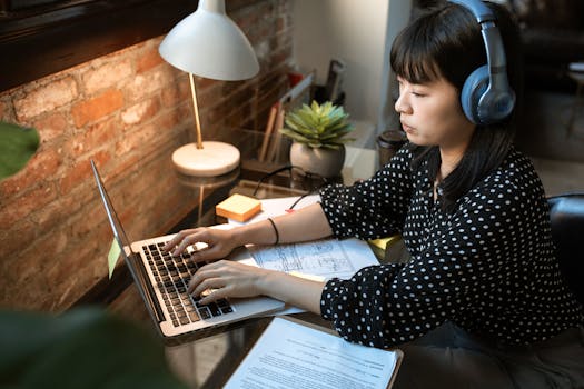 Focused woman wearing headphones, working on a laptop in a cozy home office setup with natural light.