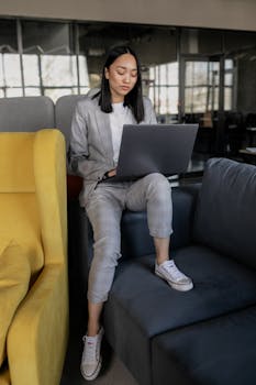 Woman in a stylish suit uses a laptop while sitting on a colorful sofa in a contemporary office setting.