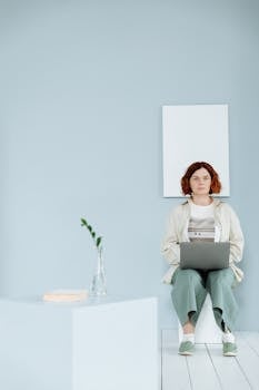 Woman with prosthetic leg sitting with laptop in minimalist room, working remotely.